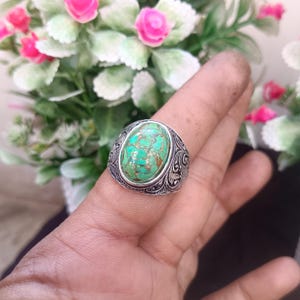 以下が含まれることがあります： 楕円形のターコイズ色の石が付いたシルバーリング。石は茶色の斑点模様です。リングのバンドには、装飾的で彫刻されたデザインが施されています。リングは指に装着されています。
