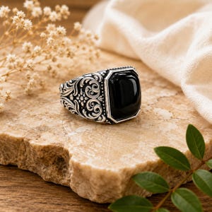 May include: A silver ring featuring a large, rectangular, polished black stone set in the centre. The band of the ring is decorated with intricate, raised scrollwork and floral patterns that extend along the sides of the metal setting.