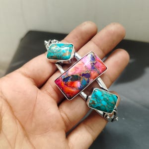 May include: A silver bracelet featuring three rectangular stones. The top and bottom stones are turquoise, while the center stone is a mix of pink, orange, and blue. The bracelet is held in a hand.