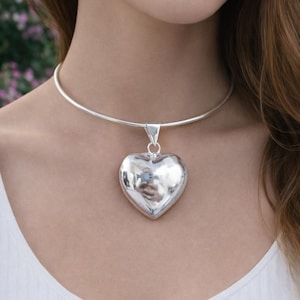 Puede incluir: Un collar plateado con un colgante en forma de corazón. El colgante cuelga de una cadena lisa y redondeada tipo gargantilla. El corazón refleja el entorno, creando un aspecto brillante. El collar es una joya.