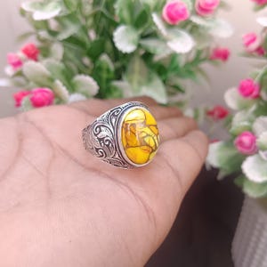 May include: Silver ring with an oval, yellow stone. The stone has a mosaic pattern with brown and yellow hues. The ring's band features an ornate, engraved design. The ring is a piece of jewellery.