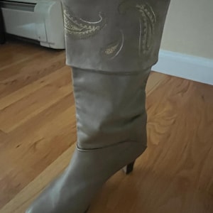 May include: A light gray mid-calf boot featuring a folded cuff at the top. The cuff is decorated with gold-toned embroidered paisley patterns. The boot has a pointed toe and a small, slender heel, constructed with a smooth, matte finish.