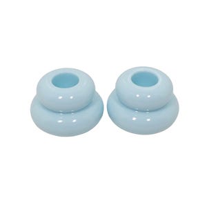 May include: Two light blue ceramic candle holders, each featuring a smooth, glossy finish and a tiered, rounded shape with a central opening designed to securely hold a standard taper candle. The objects have a uniform, solid pastel color.