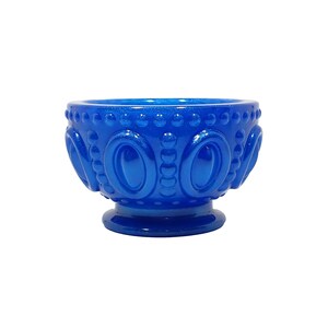 May include: A vibrant cobalt blue glass bowl featuring a raised hobnail pattern around the rim and a series of embossed oval shapes along the body. The bowl is supported by a circular pedestal base and has a glossy, reflective finish throughout.