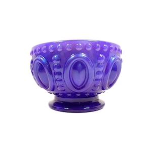 May include: A vibrant purple glass bowl with an iridescent sheen. The bowl features an ornate design with oval and circular patterns around the rim and sides. It has a pedestal base, adding to its decorative appeal. This piece is ideal for home decor.
