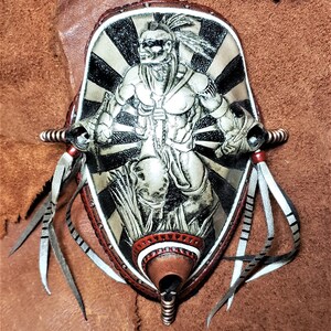 Op de afbeelding: Een leren wandhanger met een ontwerp van een indiaanse krijger. De krijger is afgebeeld in zilver met een zwart-witte achtergrond. De hanger heeft een bruine leren franje en een rode en witte rand.