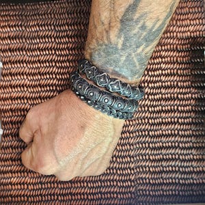 May include: Three silver-toned bracelets with intricate geometric patterns are stacked on a wrist. The bracelets have a dark, oxidized finish, and the design includes diamond and circular shapes. The background is a woven, brown surface.