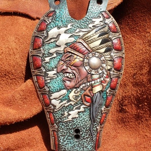 Puede incluir: Un diseño de cuero repujado de un jefe nativo americano con un tocado, pintado en colores rojo, blanco y azul. El fondo es azul turquesa con nubes blancas.