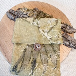 Può includere: Una piccola custodia rettangolare in tessuto con una stampa botanica di foglie nei toni del verde oliva e del marrone. La custodia presenta una patta ripiegabile fissata da un singolo bottone in legno marrone, rotondo e centrato.