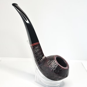 Peut inclure: Une pipe à tabac incurvée avec un foyer foncé et texturé et un tuyau noir brillant. La pipe a une bande brun rougeâtre où le tuyau rencontre le foyer. La pipe est présentée sur un support transparent sur fond blanc.