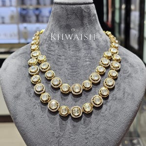Puede incluir: Un collar de oro de doble cadena con eslabones circulares que contienen piedras redondas transparentes rodeadas de pequeñas piedras blancas. El collar se exhibe sobre un busto de terciopelo gris e incluye el texto KHWAISH en letras blancas.