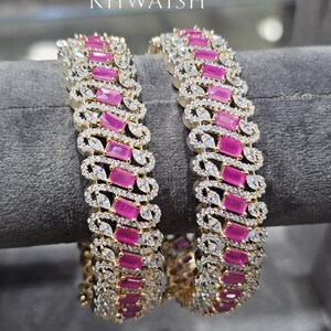 May include: Two gold bangles featuring alternating rectangular pink and clear gemstones. The bangles are displayed on a grey surface. The brand name "KHWAISH" is visible at the top.