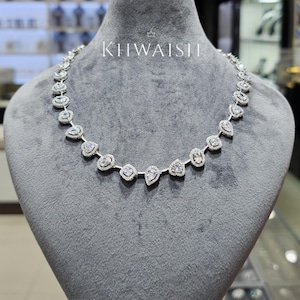 Puede incluir: Un collar de tono plateado que presenta una serie de piedras transparentes alternas en forma de pera y redondas, cada una rodeada por un halo de piedras más pequeñas en pavé. El collar muestra el texto KIIWAISHI con un icono de corona.