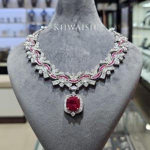 Op de afbeelding: Een zilverkleurige ketting met een centrale kussengeslepen rode edelsteen hanger omringd door kleine witte stenen. De ketting bestaat uit afwisselende segmenten van witte baguette-geslepen stenen en gebogen rijen rode stenen. Tekst leest KHWAISH.