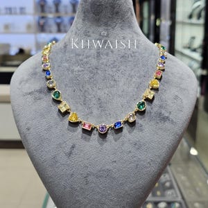 Puede incluir: Un collar de tono dorado que presenta una serie de piedras preciosas multicolores de diversas formas, incluyendo cortes redondos, cuadrados y rectangulares en tonos azul, verde, amarillo, rosa y morado. El texto KIIWAISHI aparece en blanco.
