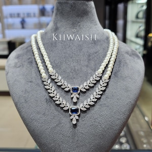 May include: Two-strand pearl necklace with diamond and sapphire pendants. The pendants feature a square sapphire center stone surrounded by a diamond leaf design. The necklaces are displayed on a gray mannequin with the text "KHWAISH".