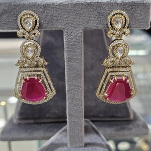 May include: A pair of gold-toned earrings with a three-tiered design. The top and middle tiers feature clear, teardrop-shaped stones surrounded by smaller, clear stones. The bottom tier showcases a large, rectangular, ruby-red stone.