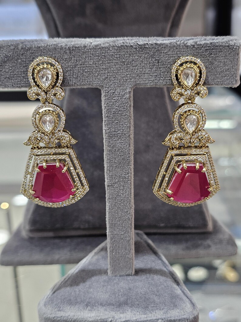 May include: A pair of gold-toned earrings with a three-tiered design. The top and middle tiers feature clear, teardrop-shaped stones surrounded by smaller, clear stones. The bottom tier showcases a large, rectangular, ruby-red stone.