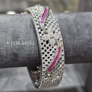 Pode incluir: Uma pulseira prateada com um design floral e incrustada com numerosas pedras pequenas e transparentes. A pulseira apresenta um motivo floral central e duas secções curvas com pedras rosa retangulares. A palavra "KIIWAISH" é visível.