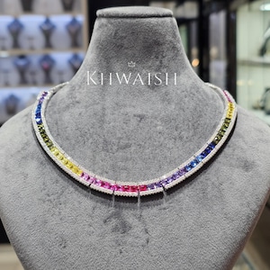 Puede incluir: Un collar que presenta una fila continua de piedras preciosas cuadradas de varios colores en un degradado de arcoíris, incluyendo amarillo, rosa, rojo, morado y azul, engastadas en un marco de metal plateado con pequeñas piedras claras y el texto KHWAISH.