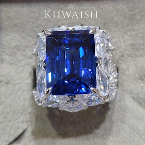 Peut inclure: Une bague ornée d'une grande pierre précieuse rectangulaire bleu foncé, entourée de petites pierres claires. Le texte "KHWAISH" est visible en haut. Le sertissage est probablement en argent ou en or blanc, mettant en valeur la pierre bleue.