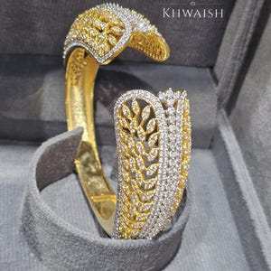 Könnte beinhalten: Ein goldfarbenes Armband in offenem Design mit zwei ausgestellten Enden. Die Oberfläche ist mit komplizierten blattförmigen Mustern verziert, die mit zahlreichen kleinen, klaren, funkelnden Steinen besetzt sind. Der Text KHWAISH ist im Hintergrund zu sehen.