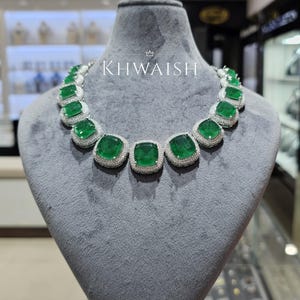 Puede incluir: Un collar de lujo con grandes esmeraldas cuadradas engastadas en un halo de pequeñas piedras brillantes. El collar se exhibe en un maniquí gris, con el nombre de la marca "KHWAISH" en texto blanco.