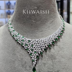 Könnte beinhalten: Eine silberfarbene Halskette mit einem blattartigen Design, das mit klaren Pavé-Steinen besetzt und mit birnenförmigen grünen Steinen akzentuiert ist. Das Schmuckstück zeigt den Text KHWAISH mit einer kleinen Krone darüber, mittig platziert.