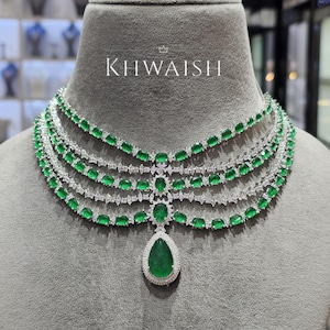 Puede incluir: Un collar de varias hebras con piedras preciosas de color verde esmeralda y piedras claras de acento. El collar tiene un colgante en forma de lágrima. El nombre de la marca "KHWAISH" se muestra en texto blanco en la parte superior de la imagen.