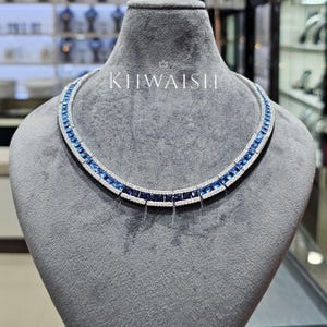 Op de afbeelding: Een ketting met een rij vierkant geslepen blauwe edelstenen geflankeerd door twee rijen kleine witte steentjes. Het sieraad wordt getoond op een grijze buste en bevat de tekst KHWAISHI in het wit boven het midden van de ketting.