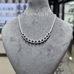 May include: A silver-toned necklace featuring a row of square-cut blue gemstones, each surrounded by a halo of small clear stones. The necklace displays the text KHWAISH in white lettering above the center of the chain on the display stand.