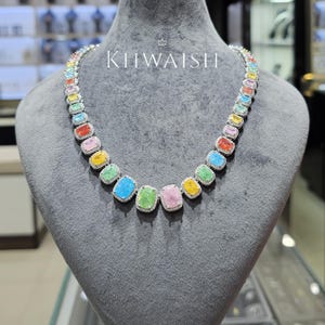 Puede incluir: Un collar que presenta una serie de piedras preciosas rectangulares multicolores en tonos rosa, amarillo, verde, azul y naranja. Cada piedra está rodeada por un borde de pequeñas piedras transparentes engastadas. La palabra KIIWAISH está impresa en blanco.