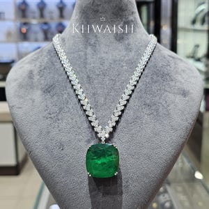 Puede incluir: Un collar con un gran colgante de piedra preciosa verde de corte cojín, suspendido de una cadena en forma de V engastada con numerosas piedras pequeñas, claras y brillantes. La palabra KHWAISH está impresa en blanco sobre el collar.
