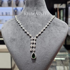 May include: A silver-toned necklace featuring a series of small, clear, sparkling stones arranged in a leaf-like pattern. The centre drop includes a prominent, pear-shaped, deep green gemstone surrounded by clear stones. The text KHWAISH is visible.