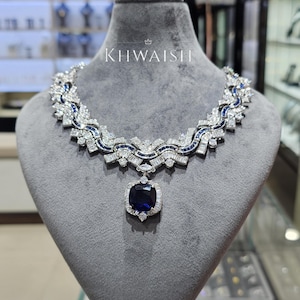 Puede incluir: Un collar de tono plateado con una gran piedra preciosa central de color azul profundo y corte cuadrado, rodeada de piedras claras más pequeñas. La cadena presenta eslabones geométricos complejos con filas de piedras claras corte baguette y detalles azules. El texto dice KHWAISHI.