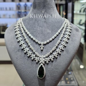 May include: A multi-strand silver-toned necklace featuring clear pavé-set stones arranged in floral patterns, with a large, pear-shaped dark green gemstone pendant at the centre. The text KHWAISH is printed in white above the necklace.