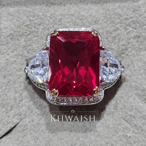 Puede incluir: Un llamativo anillo con una gran gema rectangular de color rojo rubí, flanqueada por dos piedras triangulares transparentes. La banda plateada está adornada con pequeñas piedras de acento transparentes y detalles dorados. El texto "KHWAISH" es visible en la base del anillo.