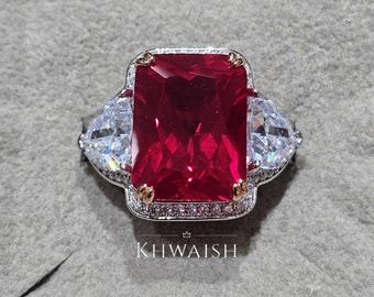 May include: A large, rectangular, ruby-red gemstone ring with two triangular, clear gemstones on either side. The ring has a silver band with small, clear accent stones and gold-colored accents. The text "KHWAISH" is at the bottom of the ring.