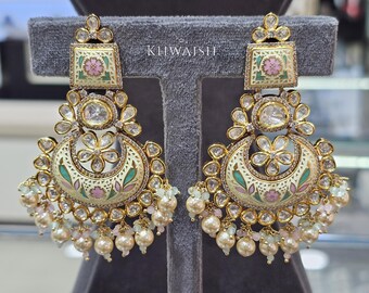 May include: Ornate gold-toned earrings with a square top and a crescent-shaped bottom. The earrings feature floral and geometric designs in pink, green, and white enamel, accented with clear crystals and dangling pearls. The word "KLIWAISH" is visible.