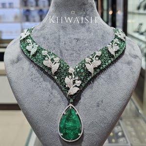 May include: A V-shaped necklace featuring numerous small green emeralds and white diamonds set in a floral pattern, with a large pear-shaped green emerald pendant at the center. The word KHWAISH is printed in white above the necklace on the display.