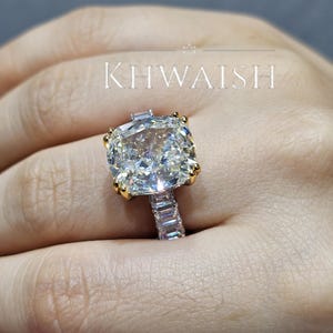 May include: A ring featuring a large cushion-cut center stone held by four yellow gold prongs, set on a band encrusted with smaller emerald-cut stones. The word KHWAISH is printed above the ring with a small crown icon centered above the text.