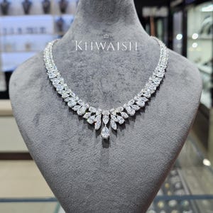 Puede incluir: Un collar de tono plateado con una serie de piedras transparentes de corte marquesa dispuestas en un diseño de doble fila, que culmina en un colgante central en forma de pera. La pieza se exhibe sobre un busto de terciopelo gris con el texto KHWAISH.