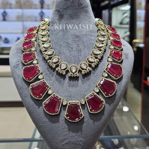 May include: A two-piece necklace set displayed on a gray mannequin. The necklaces feature large, rectangular, ruby-red gemstones set in gold-toned metal, accented with smaller clear stones. The top necklace is a choker style.