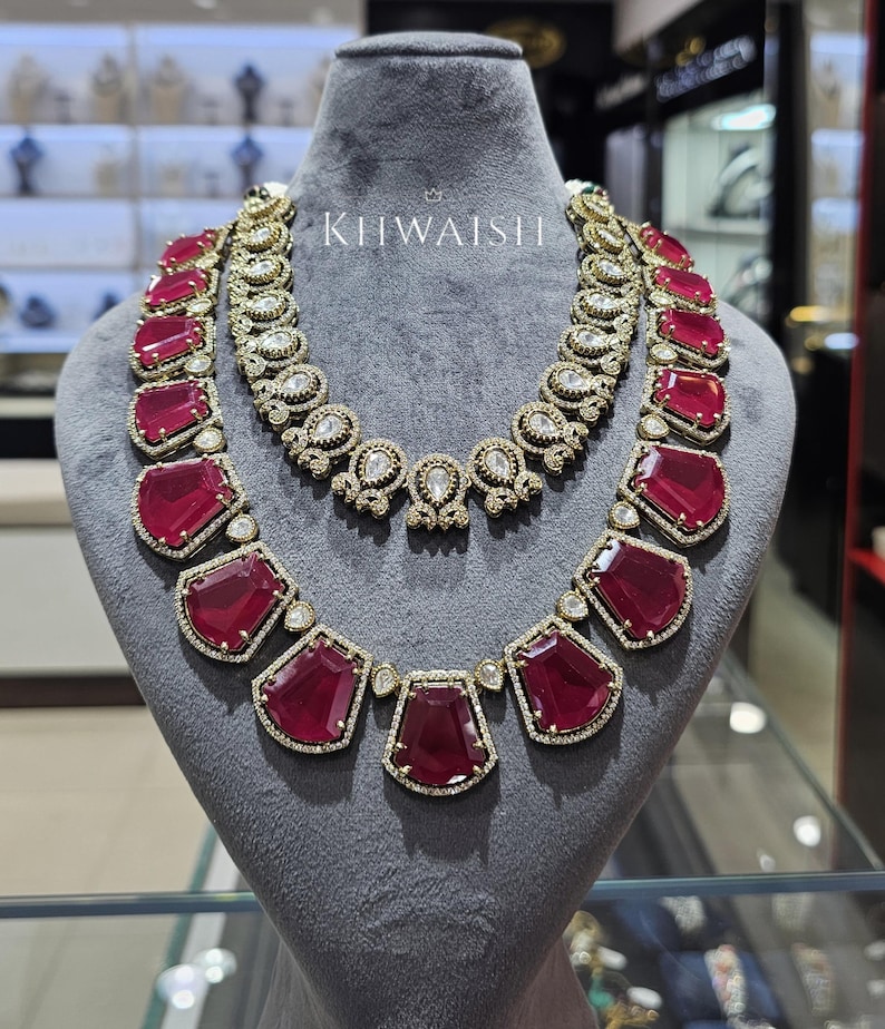 May include: A two-piece necklace set displayed on a gray mannequin. The necklaces feature large, rectangular, ruby-red gemstones set in gold-toned metal, accented with smaller clear stones. The top necklace is a choker style.