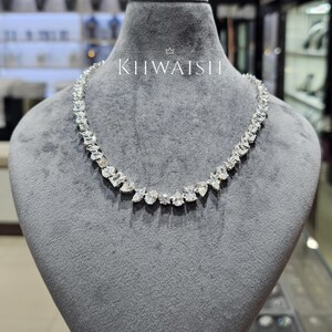 Puede incluir: Un collar de tono plateado que presenta una hilera continua de piedras transparentes facetadas en varios cortes, incluyendo formas de pera, redondas y rectangulares. El collar se exhibe sobre un busto de terciopelo gris con el texto KHWAISH en blanco.
