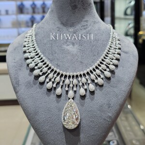 Puede incluir: Un collar de diamantes con un gran colgante en forma de pera. El collar se exhibe en un maniquí gris. El texto "KHWAISH" es visible en el maniquí. El collar está hecho de muchos diamantes pequeños.