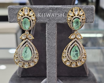 May include: A pair of gold-toned earrings with emerald-green gemstones and clear, round accent stones. The earrings have an oval top and a teardrop-shaped bottom, with the word "KHWAIISH" visible.
