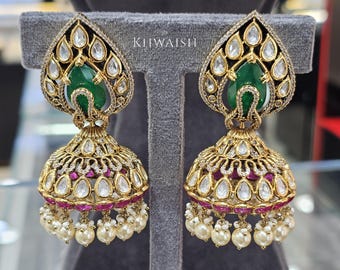 May include: A pair of gold-toned earrings with a teardrop-shaped top featuring a green gemstone and clear crystals. The lower section is bell-shaped, adorned with white crystals, red accents, and dangling pearls. The text "KHWAISH" is visible.