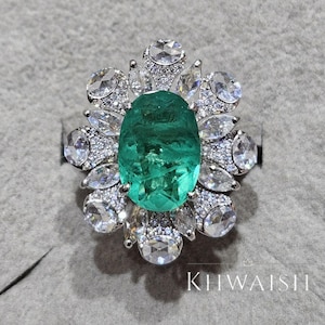 Peut inclure: Une bague ornée d'une grosse émeraude ovale au centre. L'émeraude est entourée de petites pierres claires rondes et en forme de marquise, serties dans un métal argenté ou blanc. Le texte "KHWAISH" est visible.