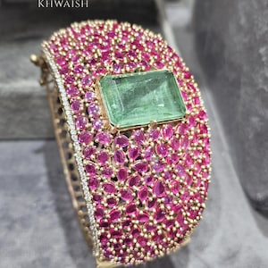 May include: A gold bracelet, densely set with pink gemstones and a central, rectangular green stone. The bracelet has a diamond-set edge and the brand name "KHWAISH". This luxury item is a bold piece of jewellery.